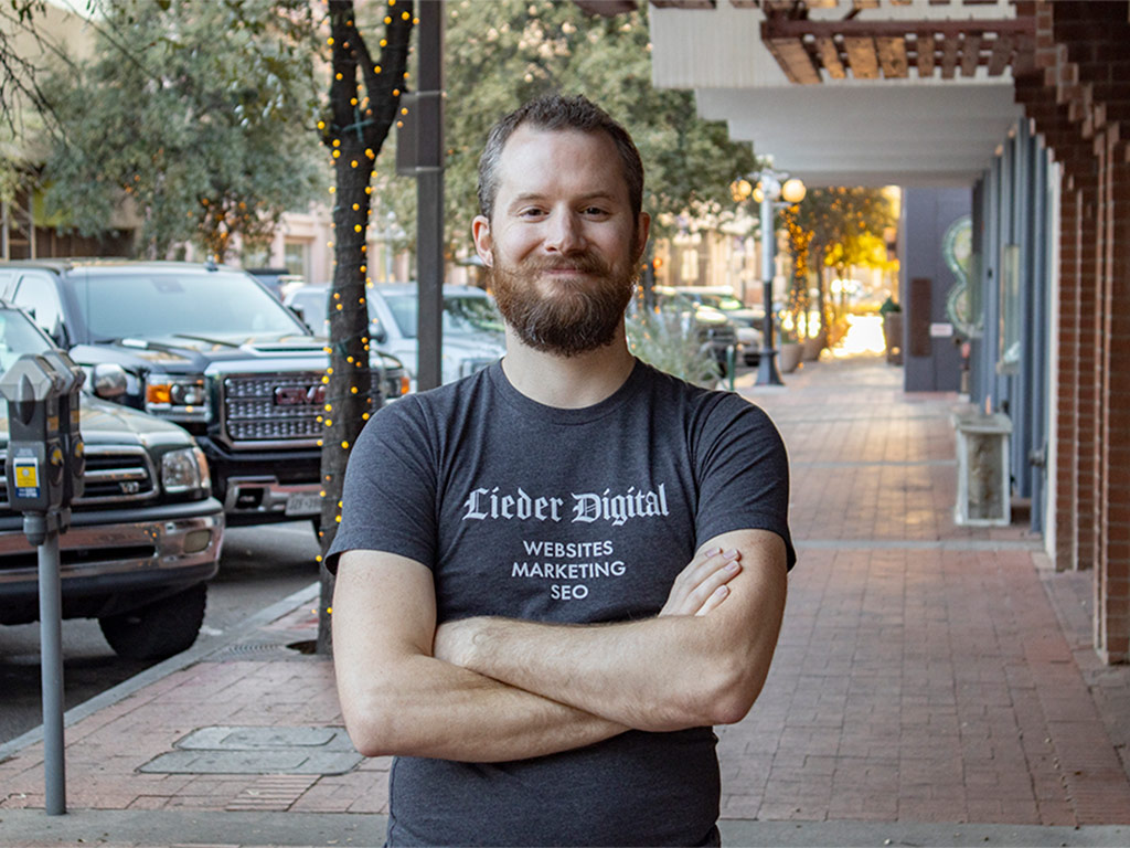 Ron Stauffer standing on the sidewalk on Pennington Street in downtown Tucson wearing a gray Lieder Digital tee shirt and smiling at the camera.